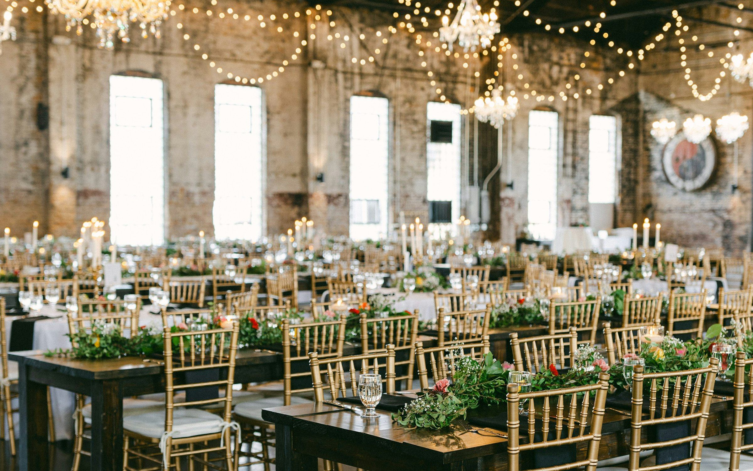 Wedding reception hall strung with fairy lights and chandeliers, tables set with gold chairs and greenery