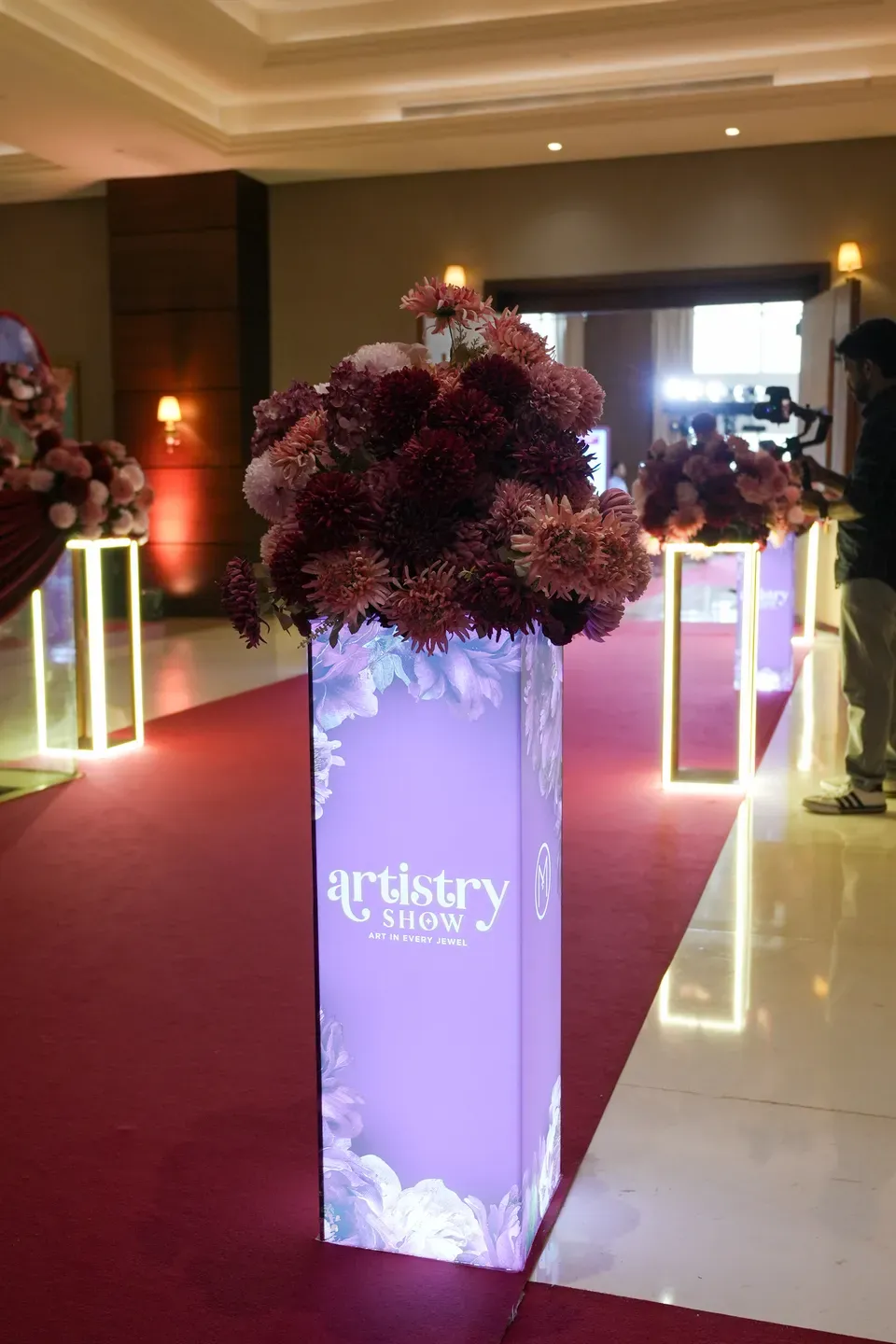 Lit branded plinth topped with burgundy dahlias on a red carpet corridor