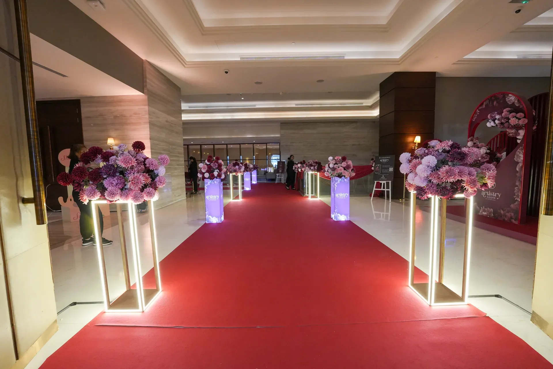 Illuminated white plinths carrying pink and burgundy florals along a red carpet corridor