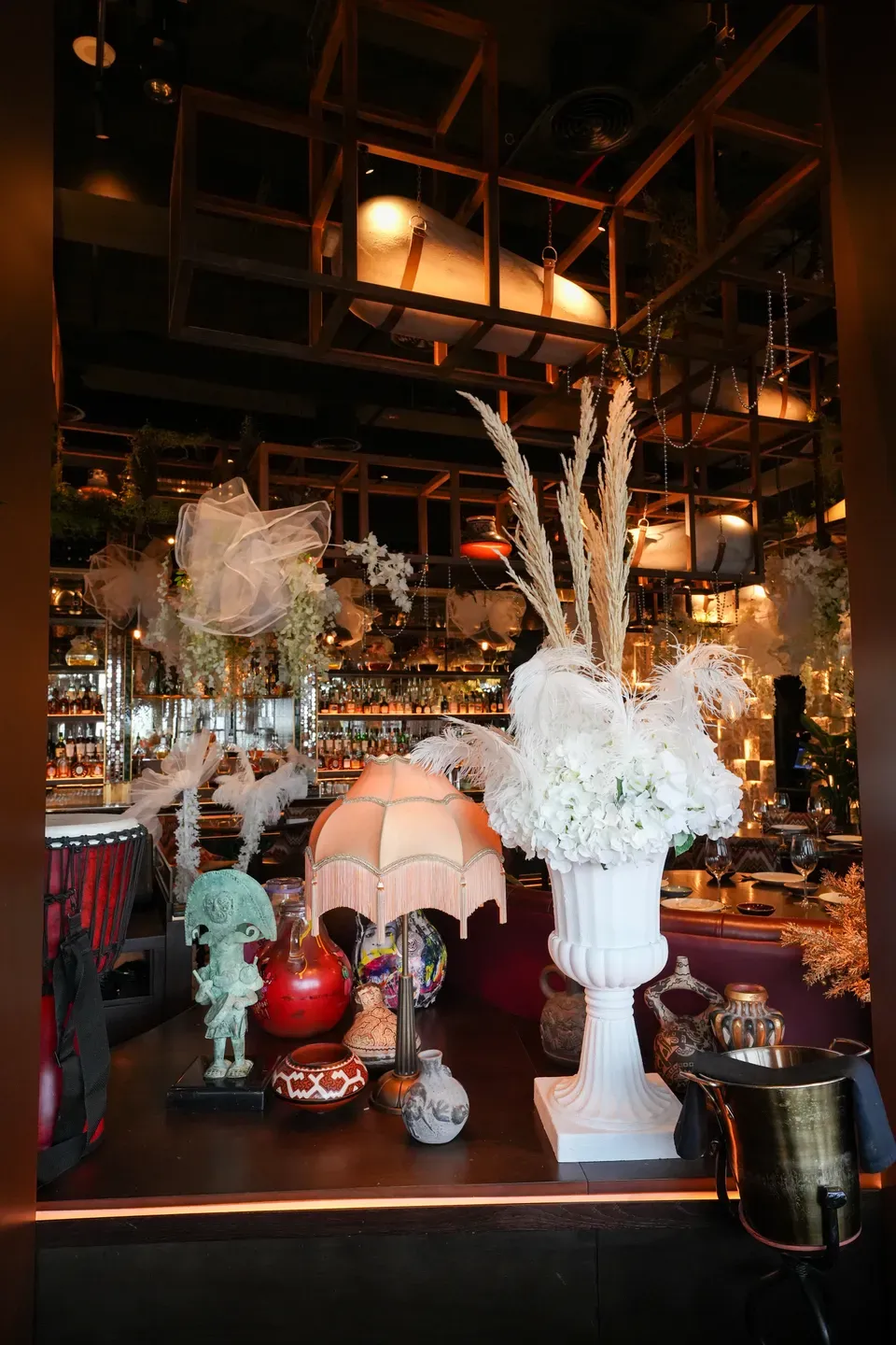 White urn of pampas and orchids beside a fringed lamp on a dressed bar counter