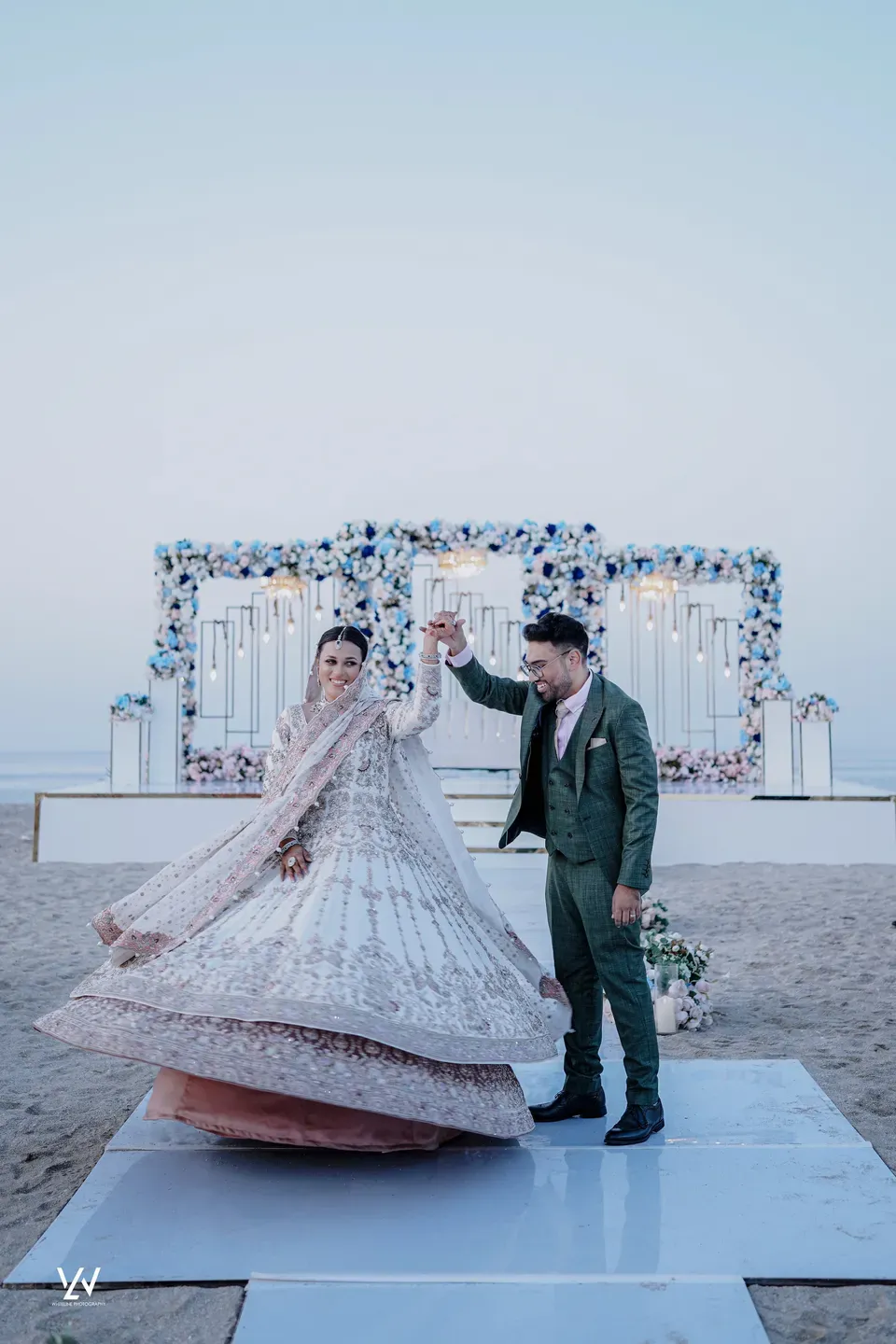 Groom turning the bride so her embroidered lehenga fans out on a beach platform at dusk