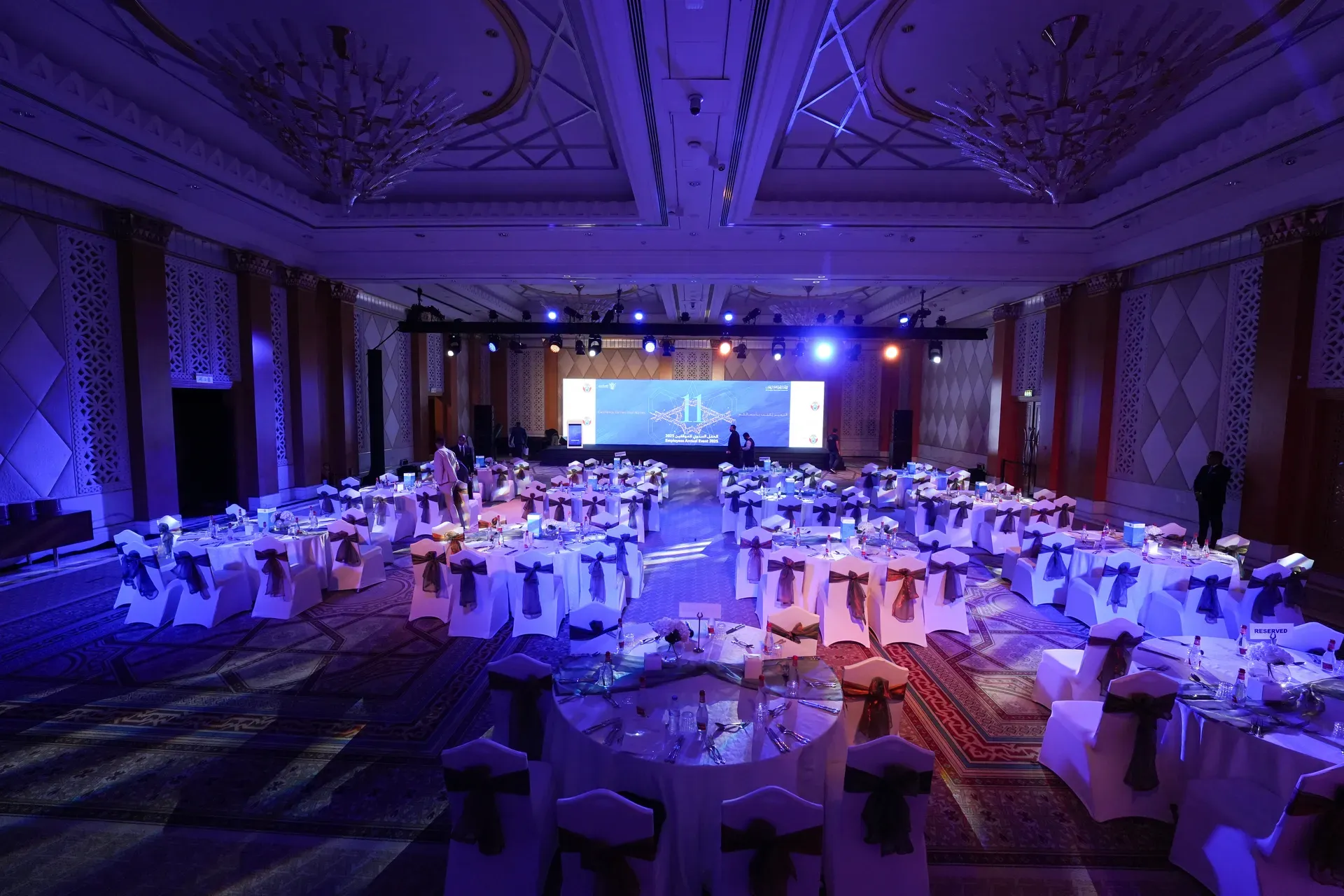 Wide view down a blue-lit ballroom towards a lit stage and screen
