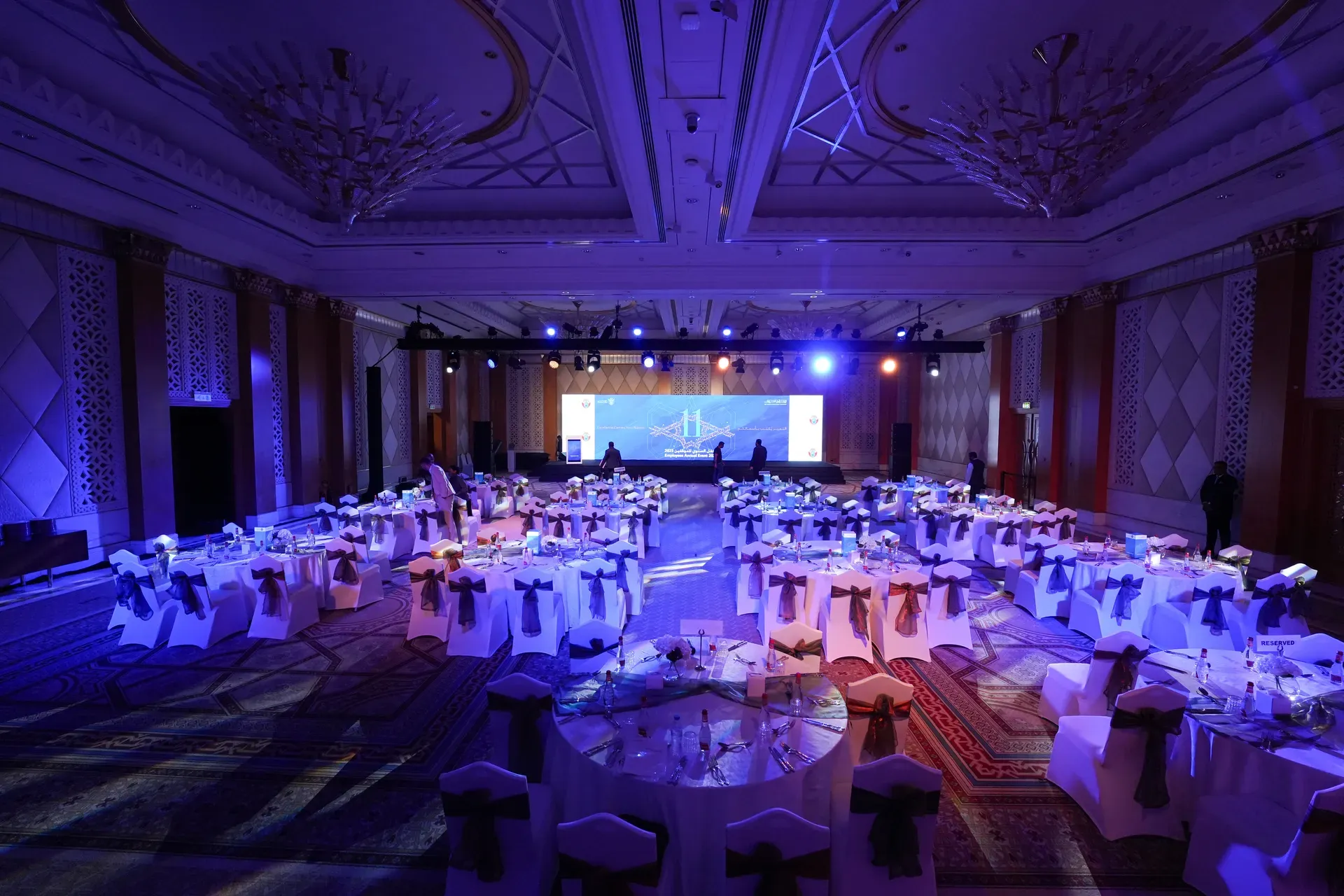 Ballroom of dressed banquet rounds under blue uplighting facing a screen
