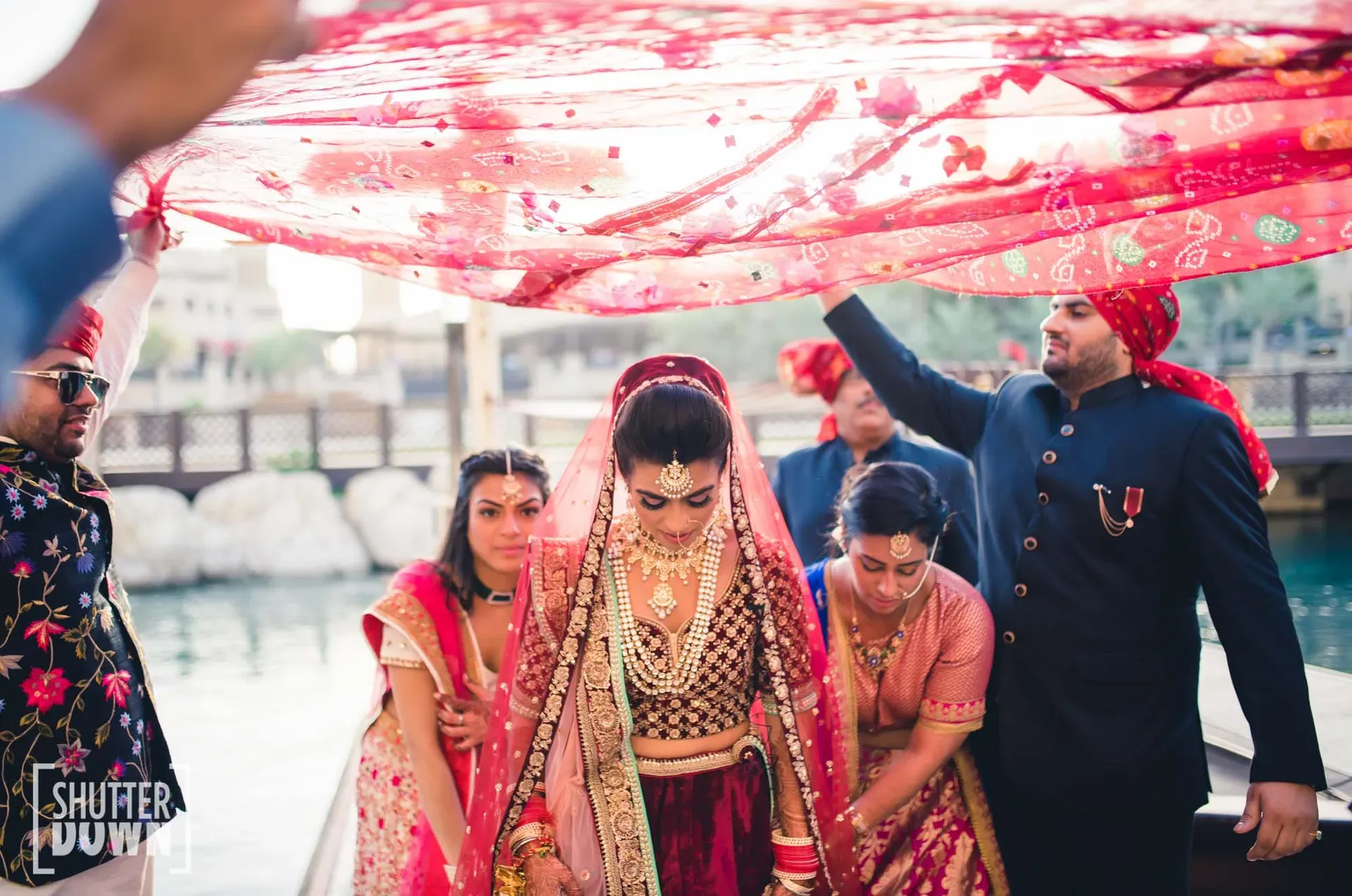Bride in red walking beneath a hand-held dupatta carried by family beside the water