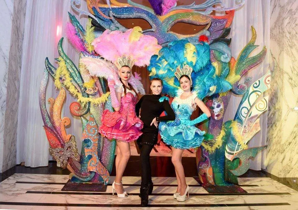 Two costumed performers flanking a guest before a painted carnival mask backdrop