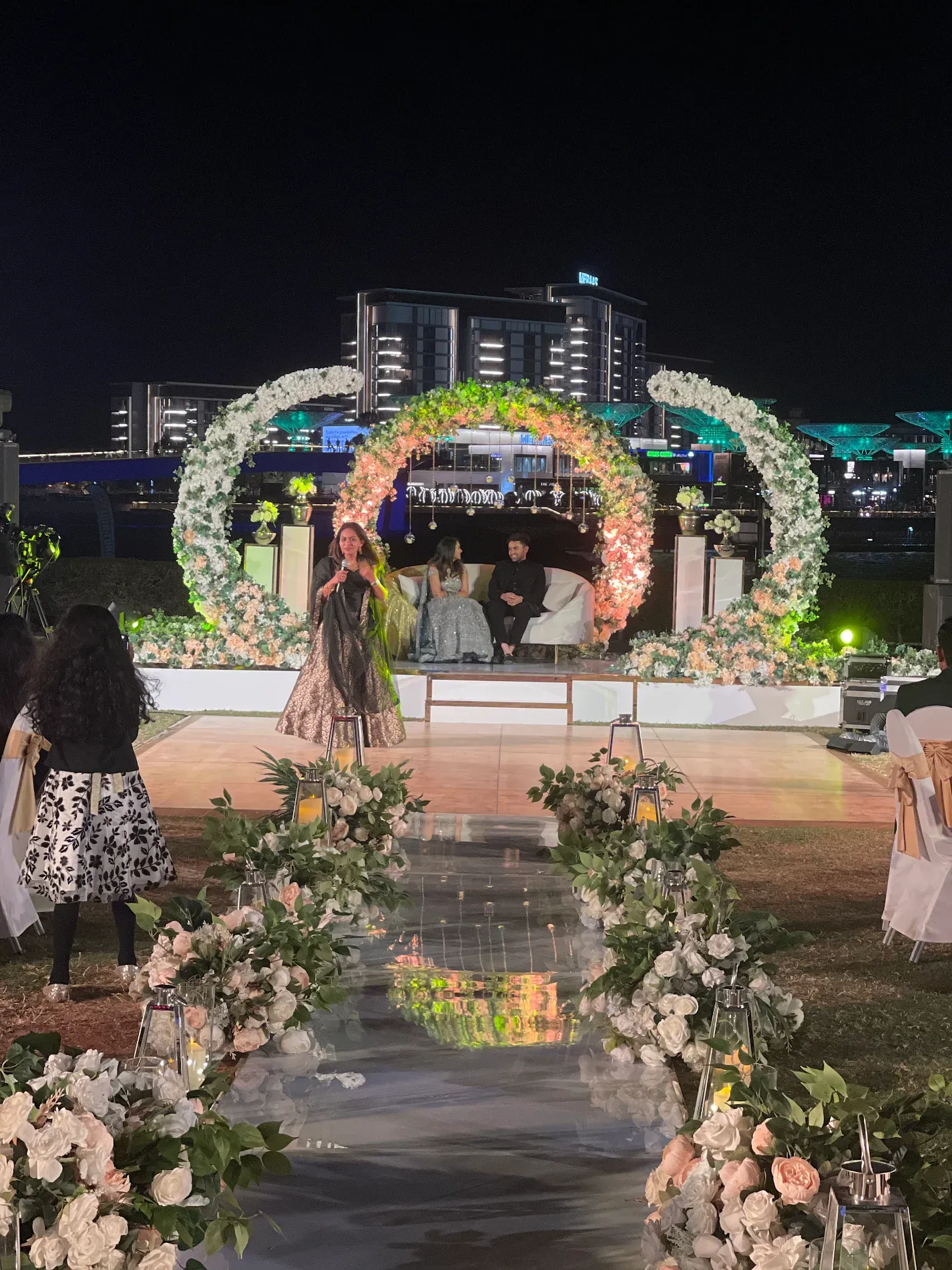 Crescent floral arch lit in peach and white above a seated couple, with the Dubai Creek Harbour skyline behind