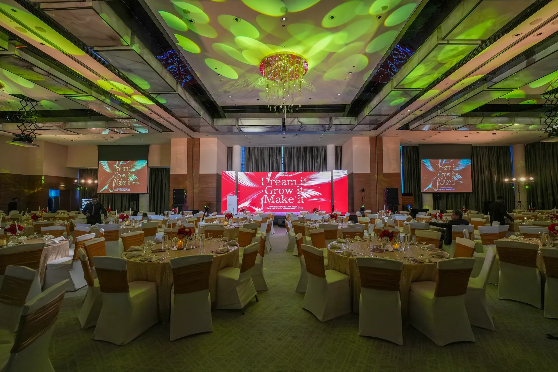Full banquet room lit green and gold beneath a central chandelier facing a red stage