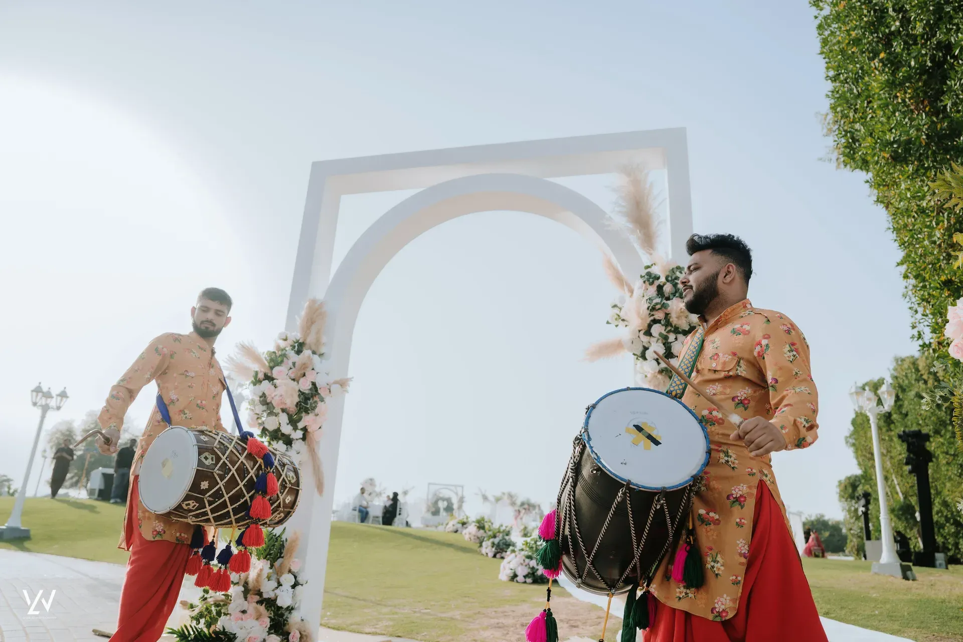 Two dhol players in orange kurtas drumming beside a white arch dressed with pampas