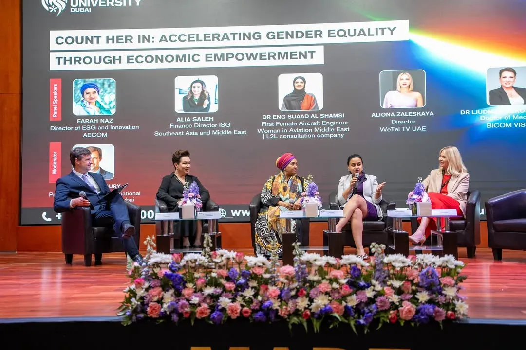 Five panellists seated on an auditorium stage below a screen of speaker portraits