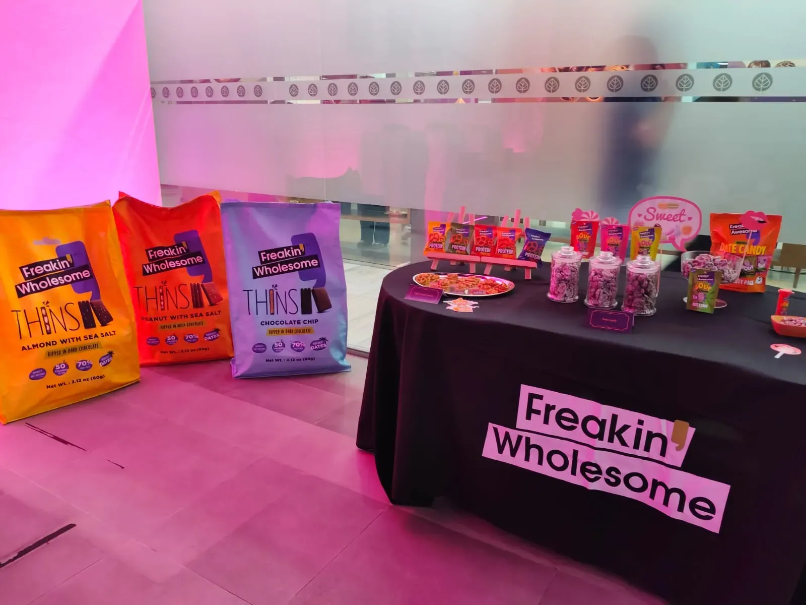 Product sampling table beside oversized snack packs under pink lighting