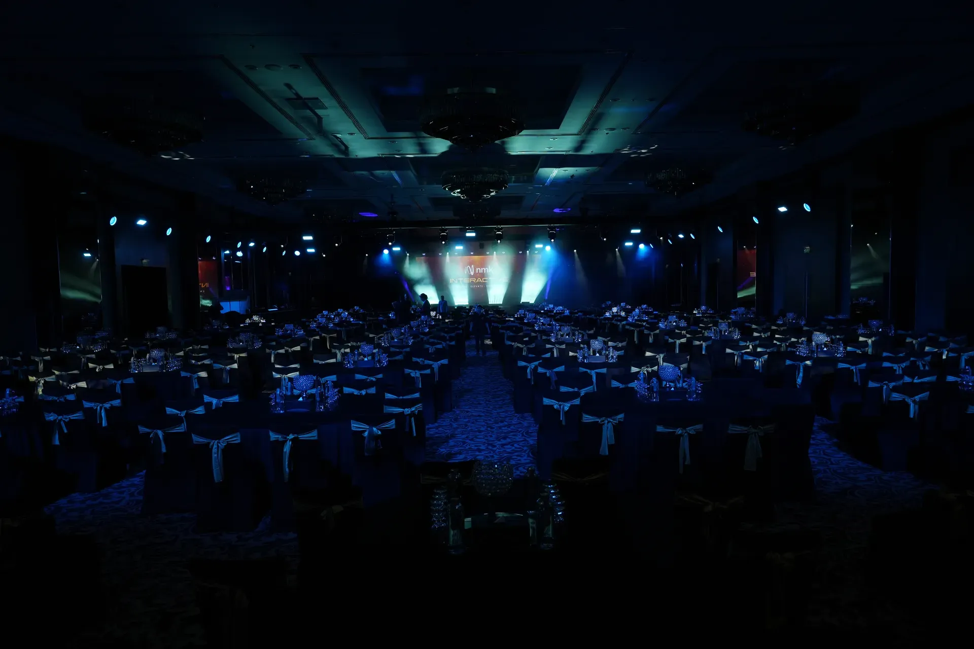 Darkened ballroom of banquet rounds facing a lit stage before guests arrive