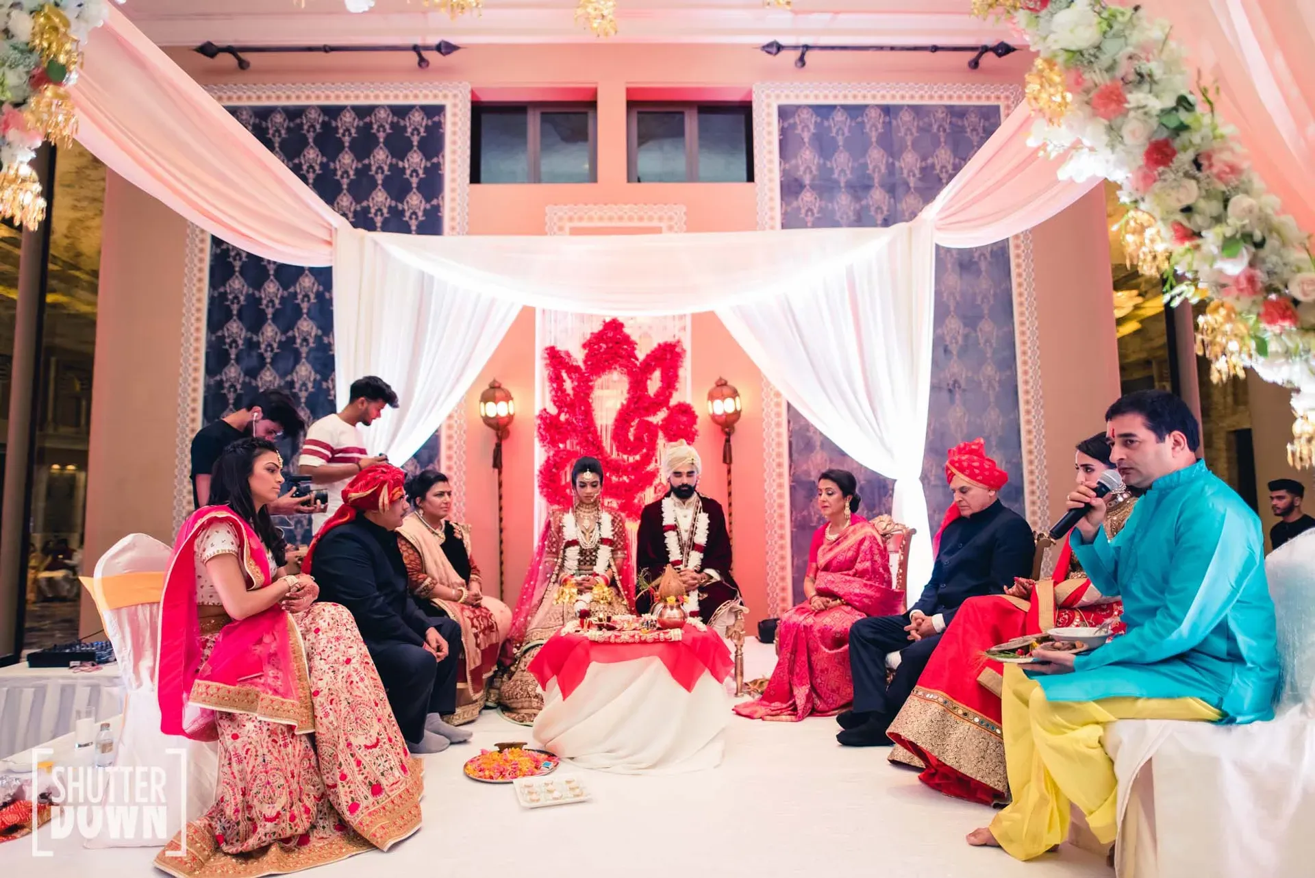 Wedding party seated on a draped mandap stage with a red Ganesh motif behind