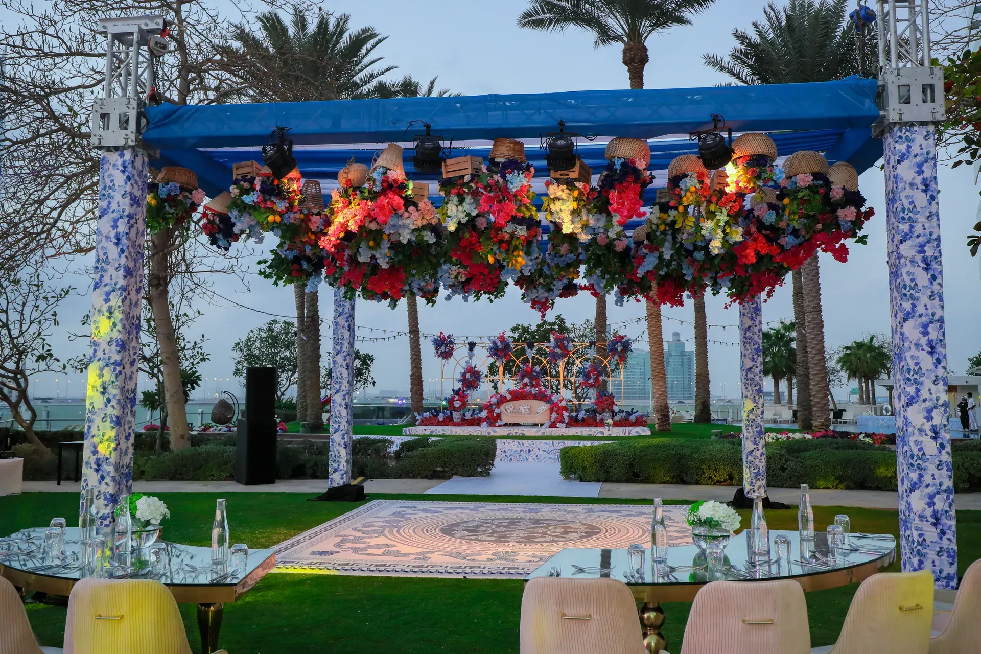 Open-air mandap on a lawn under a blue canopy massed with tropical flowers and palms