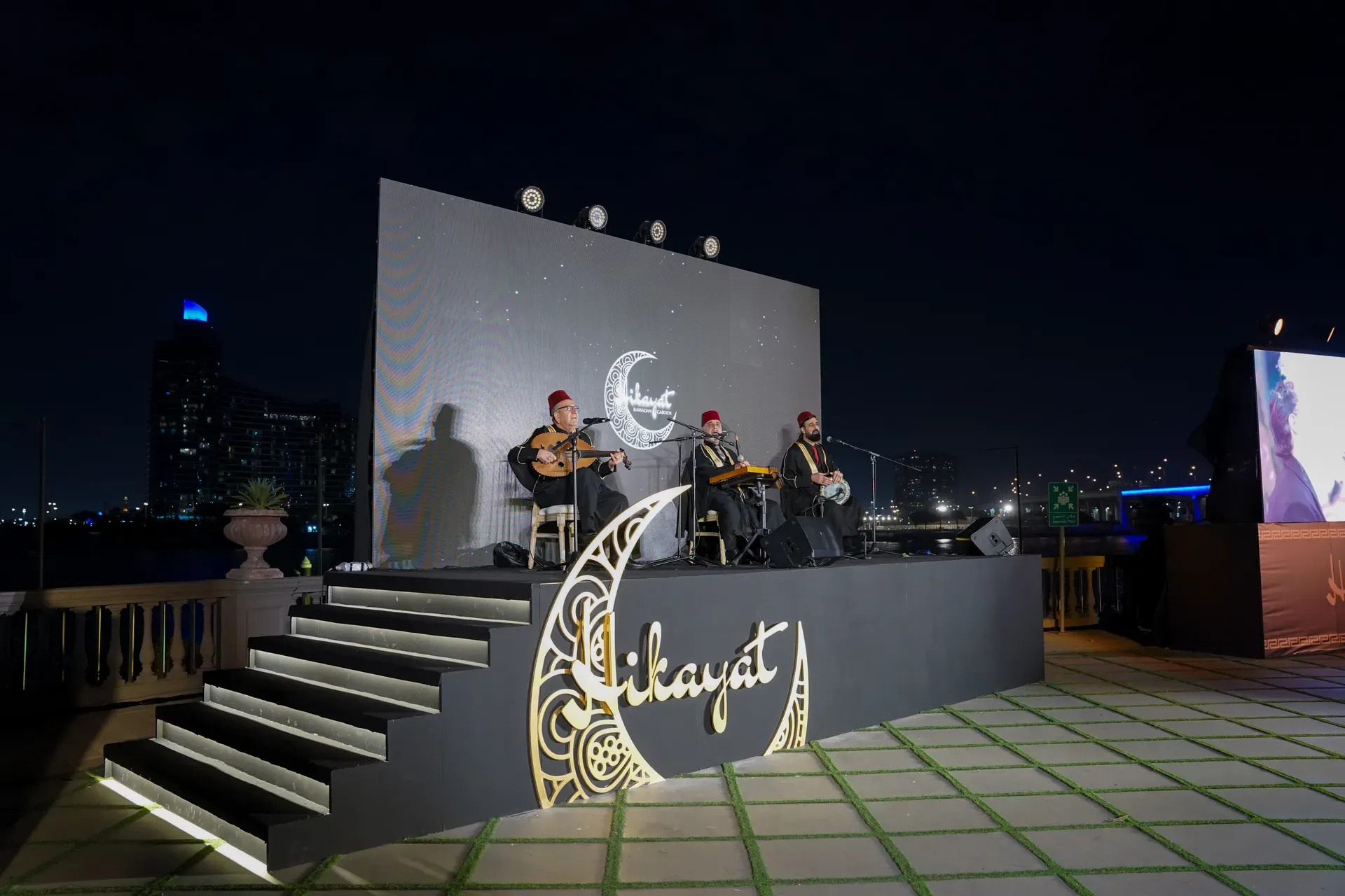 Three musicians playing on a rooftop stage below a crescent moon logo at night