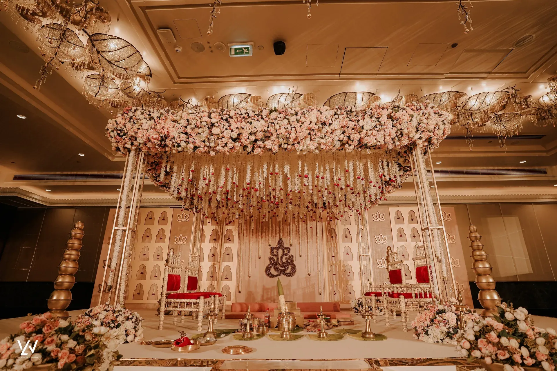 Mandap with red and gold seats beneath a dense blush floral canopy and brass urns