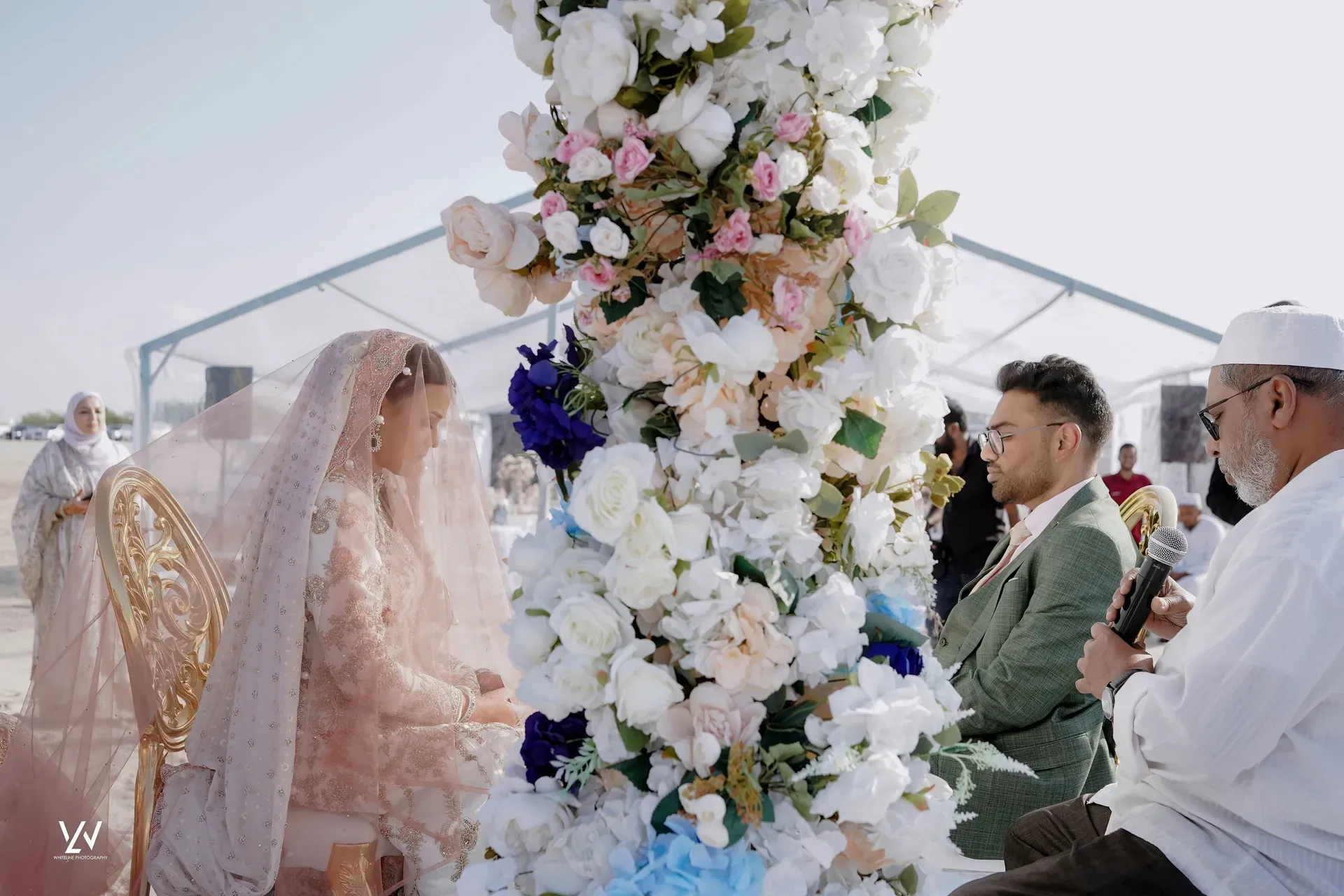 Bride and groom seated either side of a tall white and pink floral column in a marquee