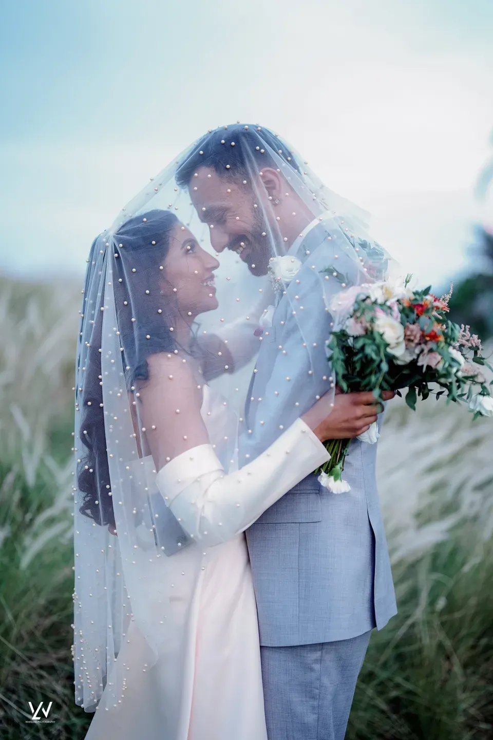 Bride and groom sharing a pearl-studded veil in a field of tall grasses
