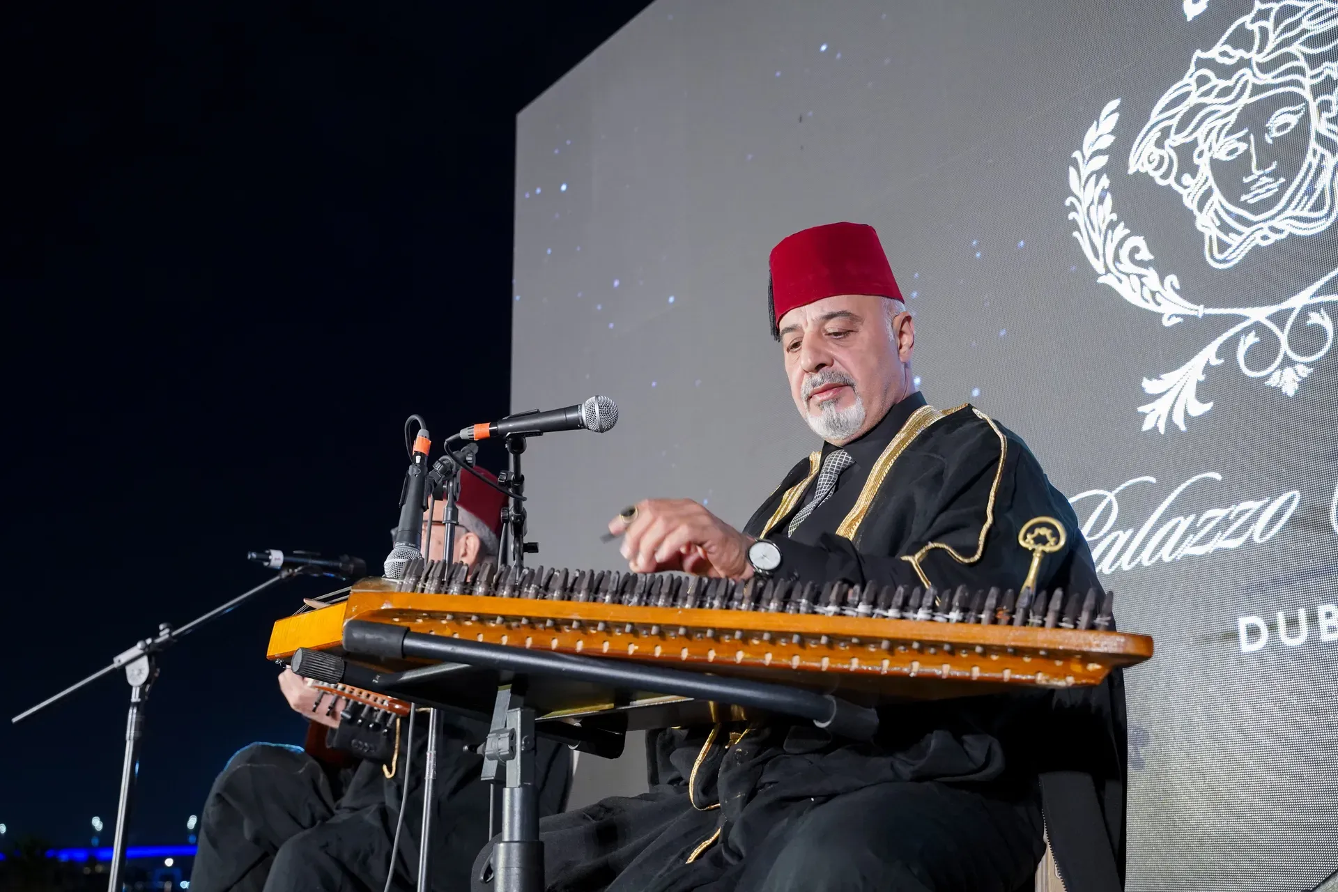 Musician in a red tarboosh and embroidered robe playing a qanun at a microphone