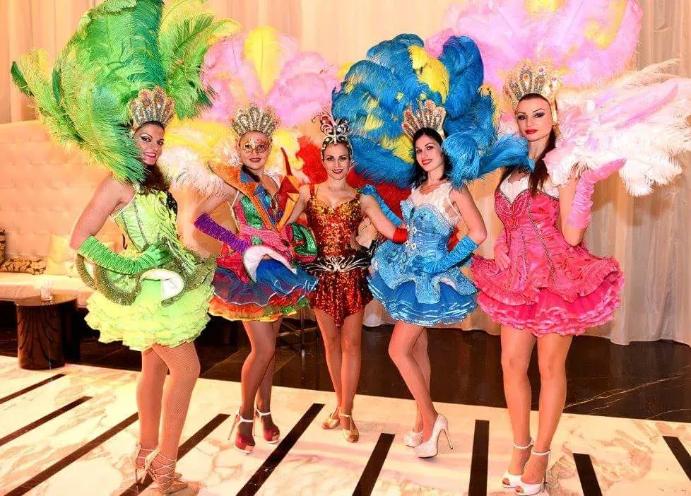 Six performers in feathered carnival costumes standing in a row on a dance floor