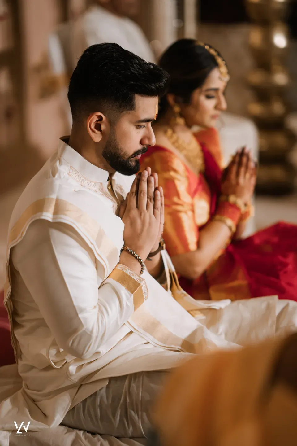 Couple in gold-bordered ivory and red silk with palms pressed together during a temple ceremony
