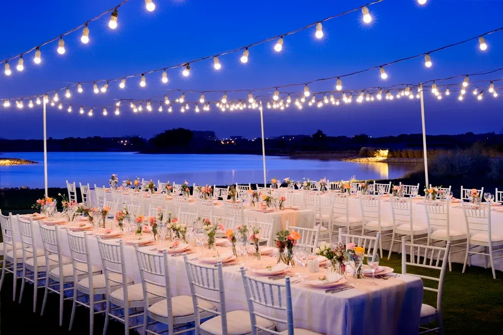 Long banquet tables under strings of festoon bulbs beside a lake at dusk