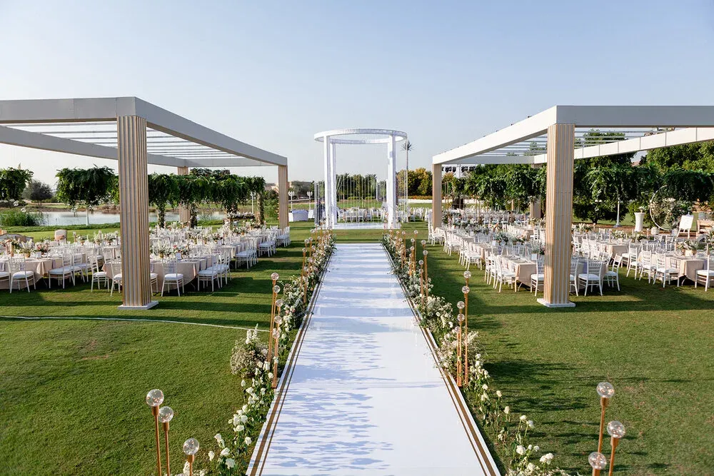 White ceremony aisle running between two pergolas of banquet tables on a green lawn