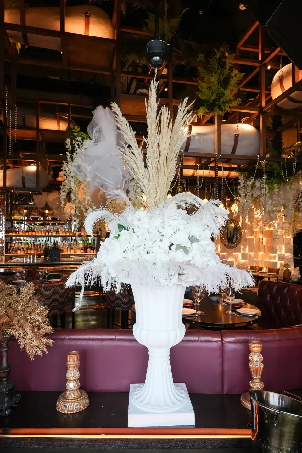 Tall white urn of pampas and white blooms in front of a purple velvet banquette