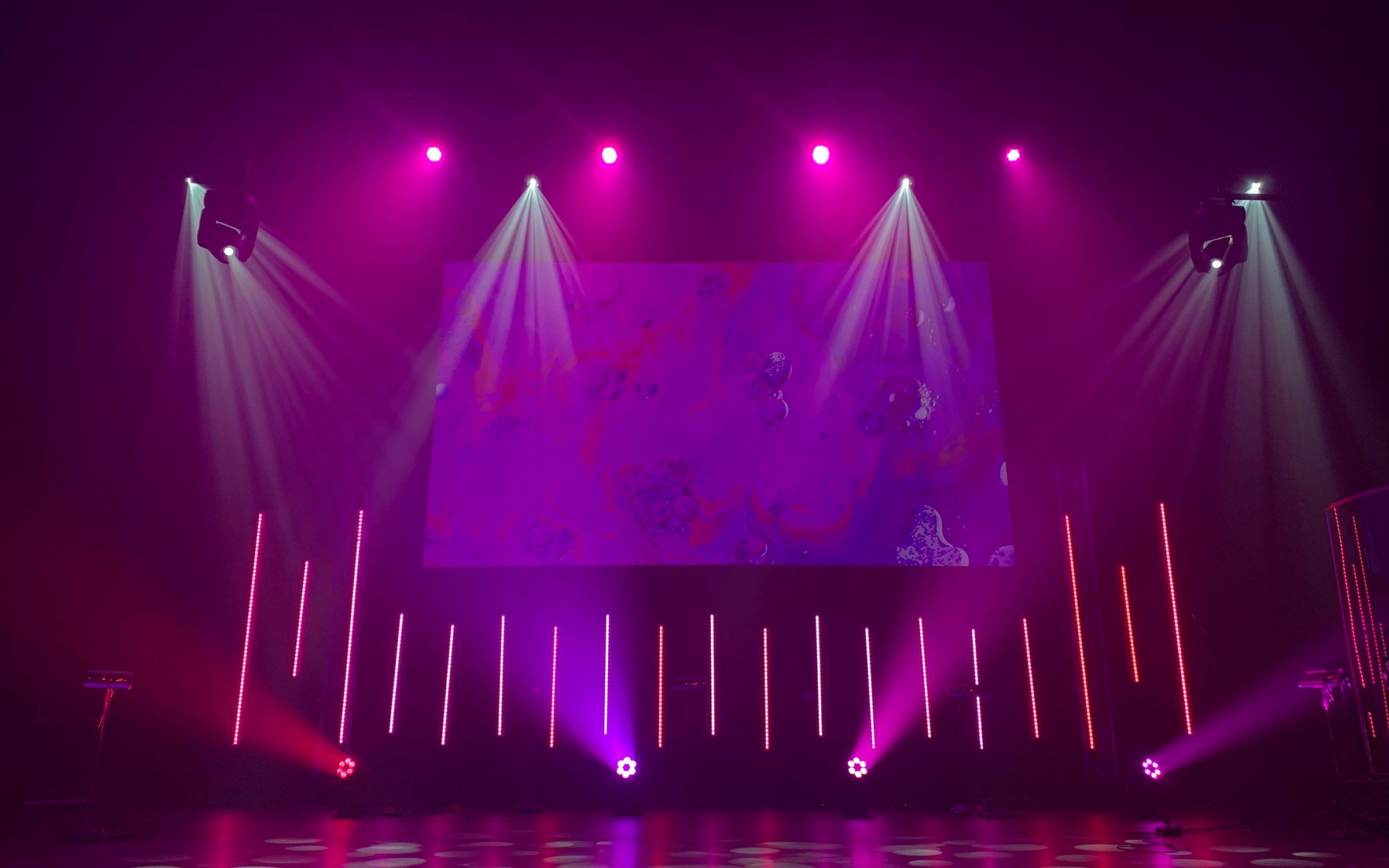 Empty stage lit with pink and purple moving-head fixtures and a blank abstract screen